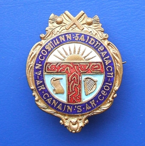 An early badge of the Scottish-speaking (Gaelic) rights movement An Comunn Gaidhealach with the Fenian Sunburst symbol as a sun rising above the horizon, Scotland